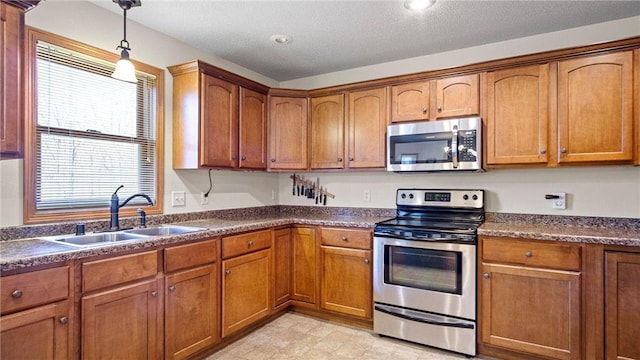 kitchen featuring brown cabinetry, a sink, stainless steel appliances, dark countertops, and decorative light fixtures