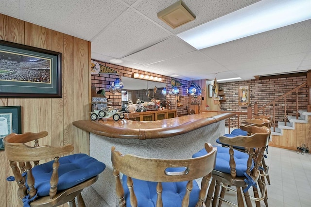 bar with a drop ceiling, wooden walls, a dry bar, and brick wall