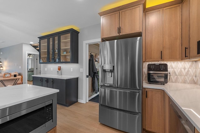 kitchen with backsplash, appliances with stainless steel finishes, brown cabinetry, light wood finished floors, and glass insert cabinets