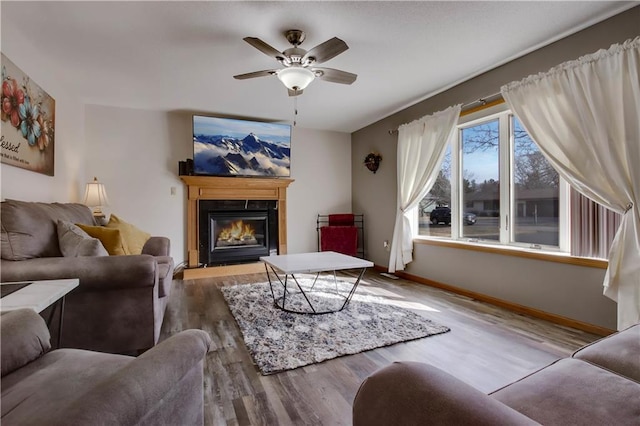 living area with a glass covered fireplace, wood finished floors, baseboards, and ceiling fan