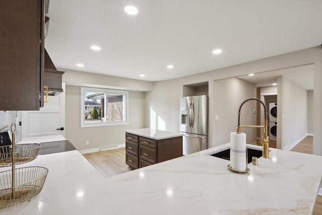 kitchen with light wood-type flooring, stainless steel fridge, dark brown cabinetry, stacked washer / dryer, and light stone countertops