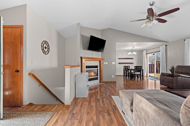 living room featuring wood finished floors, baseboards, lofted ceiling, a tile fireplace, and ceiling fan with notable chandelier