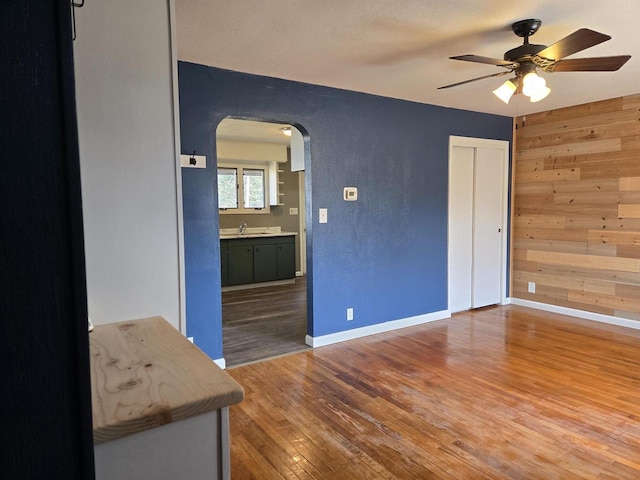 unfurnished living room featuring baseboards, ceiling fan, wood walls, wood finished floors, and arched walkways