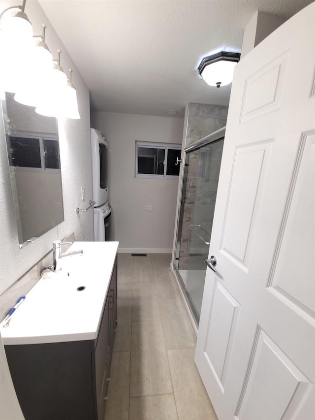 full bath featuring a stall shower, a textured ceiling, stacked washer / dryer, baseboards, and vanity