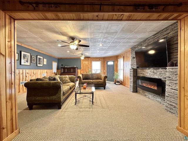 carpeted living area with wooden walls, crown molding, a stone fireplace, an ornate ceiling, and a ceiling fan