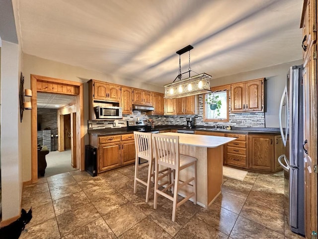 kitchen with brown cabinetry, under cabinet range hood, appliances with stainless steel finishes, backsplash, and a center island