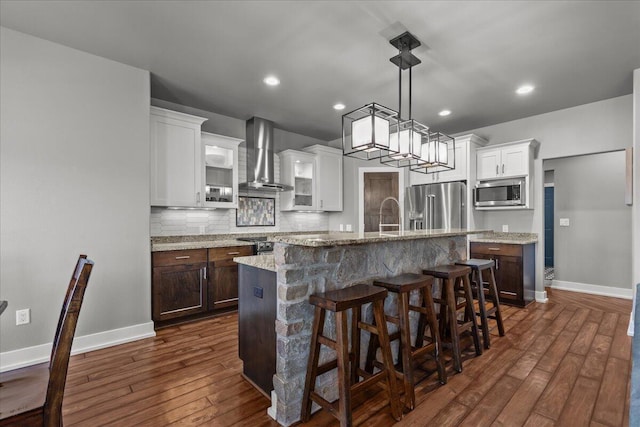 kitchen featuring stainless steel appliances, an island with sink, white cabinets, and wall chimney range hood