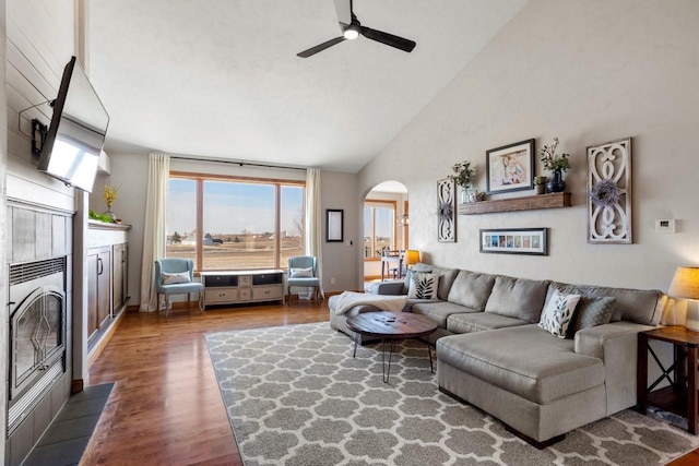 living area featuring high vaulted ceiling, a ceiling fan, a tiled fireplace, wood finished floors, and arched walkways