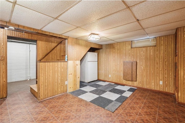 finished below grade area featuring wooden walls, baseboards, a drop ceiling, freestanding refrigerator, and tile patterned floors