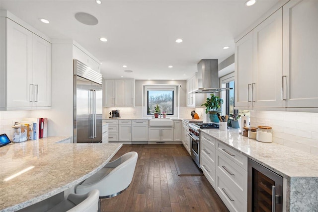 kitchen with light stone counters, premium appliances, white cabinetry, wine cooler, and wall chimney exhaust hood