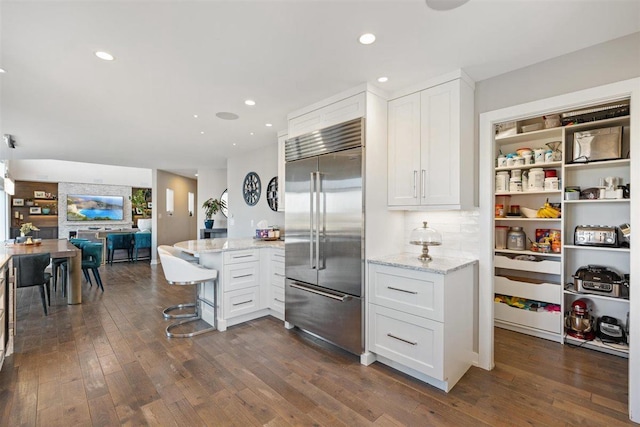 kitchen with light stone counters, a peninsula, dark wood-type flooring, built in fridge, and white cabinetry