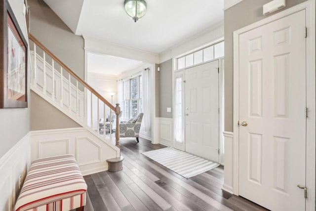 foyer featuring a wainscoted wall, stairway, crown molding, a decorative wall, and dark wood-style flooring