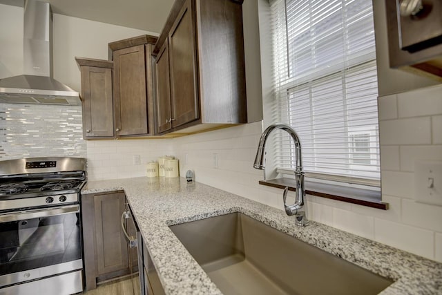 kitchen with light stone countertops, a sink, decorative backsplash, gas range, and wall chimney range hood