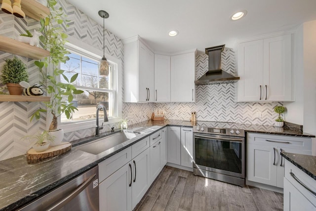 kitchen with decorative light fixtures, appliances with stainless steel finishes, white cabinetry, wall chimney exhaust hood, and a sink