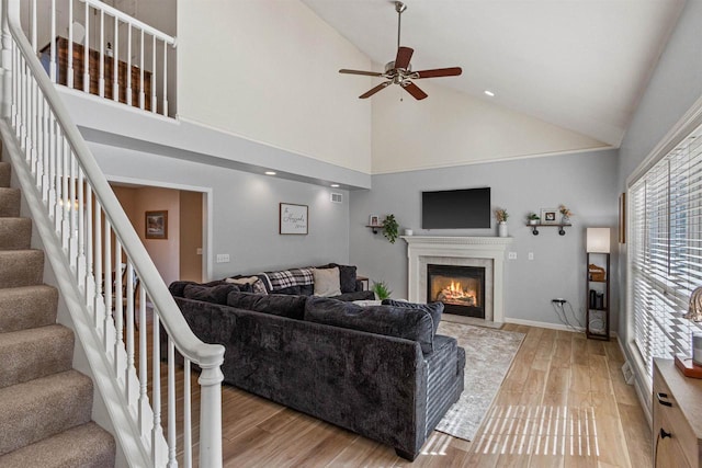 living area featuring stairs, light wood-type flooring, a glass covered fireplace, high vaulted ceiling, and a ceiling fan