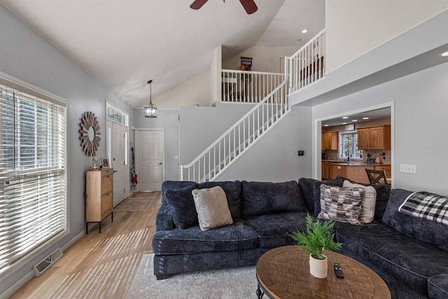 living room featuring light wood-type flooring, visible vents, high vaulted ceiling, ceiling fan, and stairs