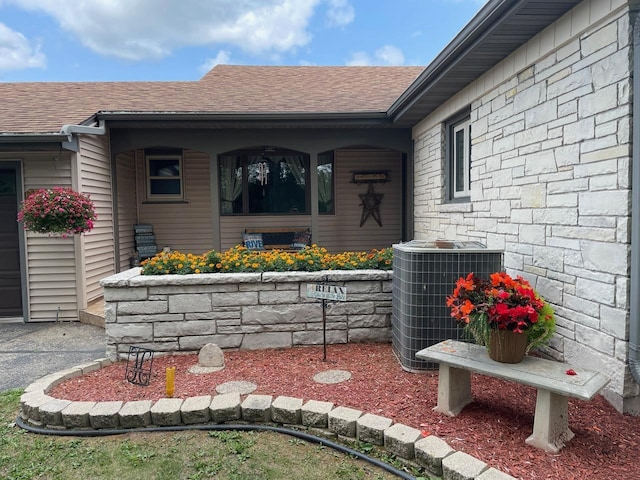 view of exterior entry featuring stone siding, central AC, and roof with shingles