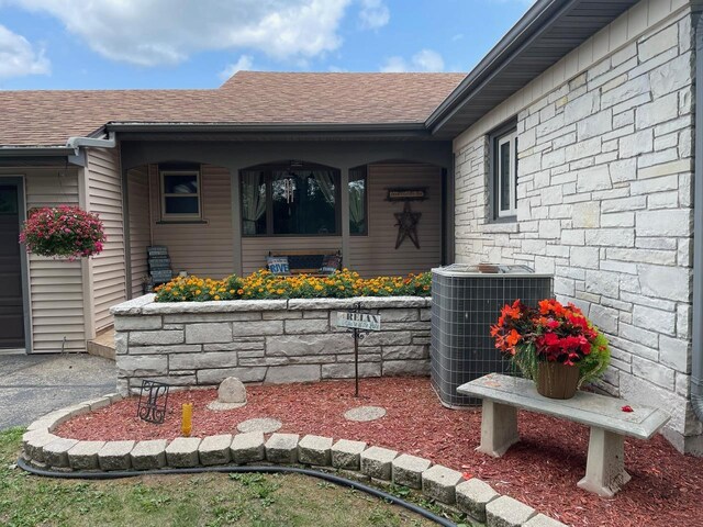 entrance to property with central air condition unit, a shingled roof, and stone siding