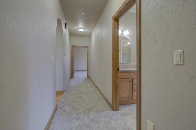 hallway featuring baseboards, arched walkways, a sink, light carpet, and a textured wall