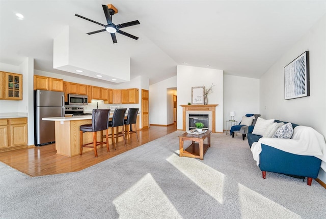 living room with a ceiling fan, high vaulted ceiling, light wood-style floors, a glass covered fireplace, and light colored carpet