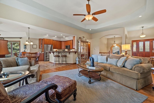 living area with a raised ceiling, recessed lighting, ceiling fan with notable chandelier, and light wood-type flooring