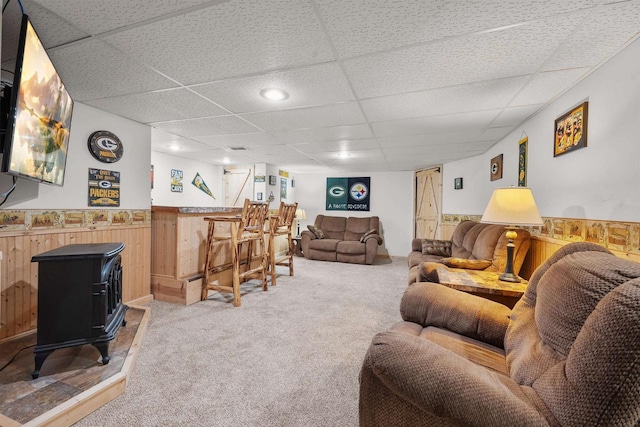 living room with a drop ceiling, wood walls, wainscoting, light colored carpet, and a wood stove