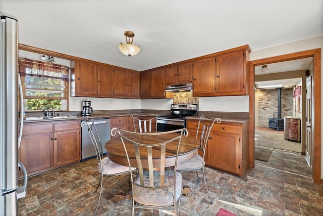 kitchen with a wood stove, stainless steel appliances, stone finish floor, under cabinet range hood, and dark countertops