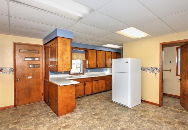 kitchen featuring a sink, freestanding refrigerator, brown cabinetry, light countertops, and a paneled ceiling