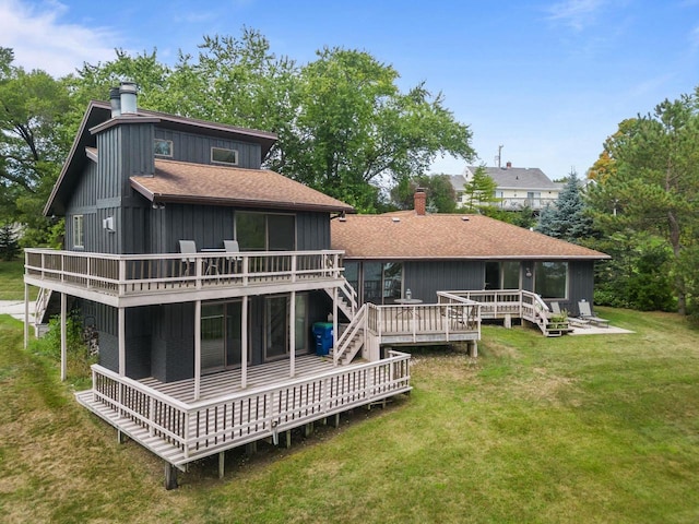 back of property with a shingled roof, a wooden deck, a lawn, and a chimney