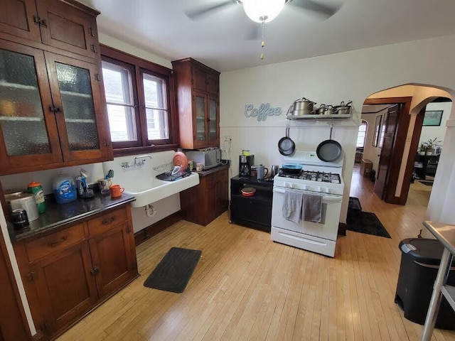 kitchen featuring a ceiling fan, light wood finished floors, white range with gas cooktop, arched walkways, and dark countertops