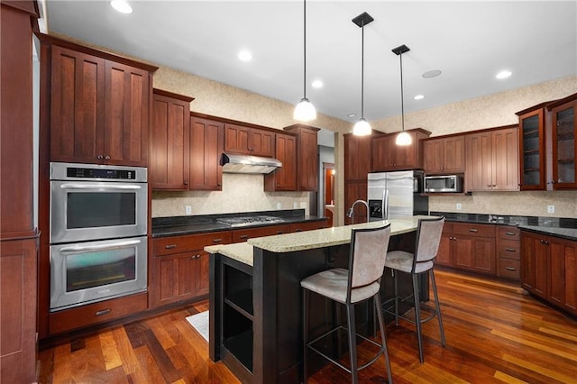 kitchen featuring dark wood-style floors, a kitchen island with sink, stainless steel appliances, under cabinet range hood, and a kitchen breakfast bar