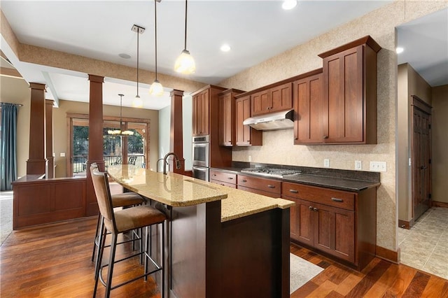 kitchen featuring under cabinet range hood, dark wood finished floors, a breakfast bar area, decorative columns, and stainless steel appliances