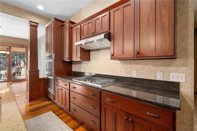 kitchen with brown cabinets, dark wood-type flooring, under cabinet range hood, stainless steel appliances, and ornate columns