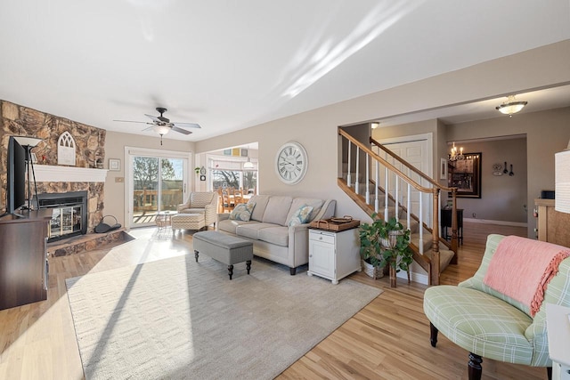 living room featuring stairway, ceiling fan with notable chandelier, a stone fireplace, and light wood-style floors