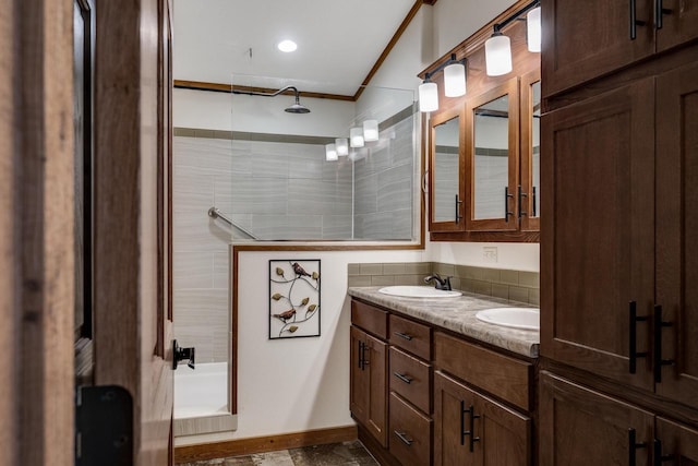 full bathroom featuring baseboards, ornamental molding, a tile shower, double vanity, and a sink