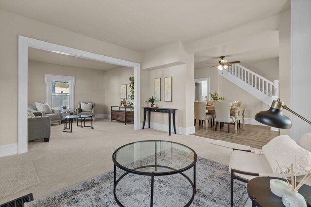 living room with stairway, a ceiling fan, visible vents, baseboards, and carpet flooring