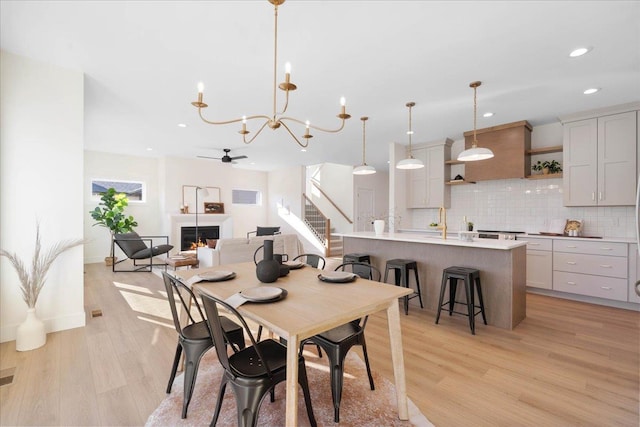 dining space featuring light wood finished floors, recessed lighting, a warm lit fireplace, stairs, and ceiling fan with notable chandelier