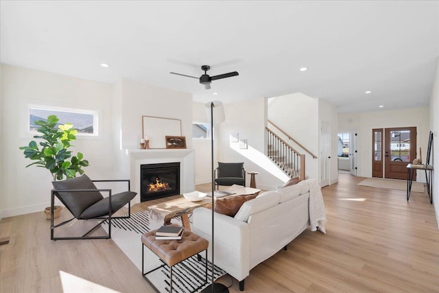 living room featuring light wood finished floors, a glass covered fireplace, stairs, and ceiling fan