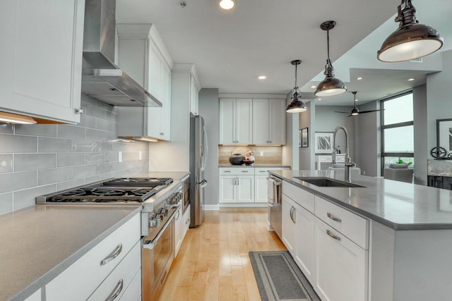 kitchen with an island with sink, a ceiling fan, a sink, stainless steel appliances, and wall chimney range hood