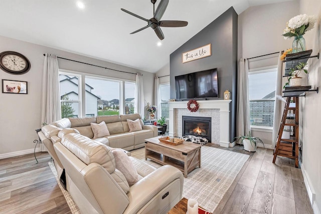 living area featuring baseboards, light wood finished floors, high vaulted ceiling, a tile fireplace, and ceiling fan