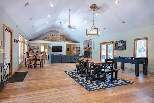 dining area featuring light wood finished floors, a stone fireplace, ceiling fan with notable chandelier, and high vaulted ceiling