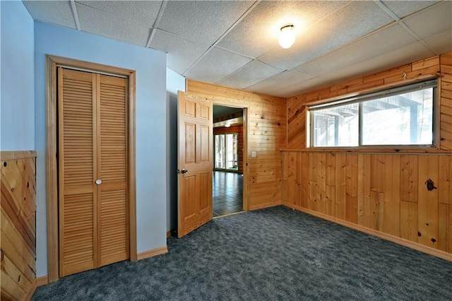 unfurnished bedroom featuring a closet, wood walls, a paneled ceiling, and dark colored carpet