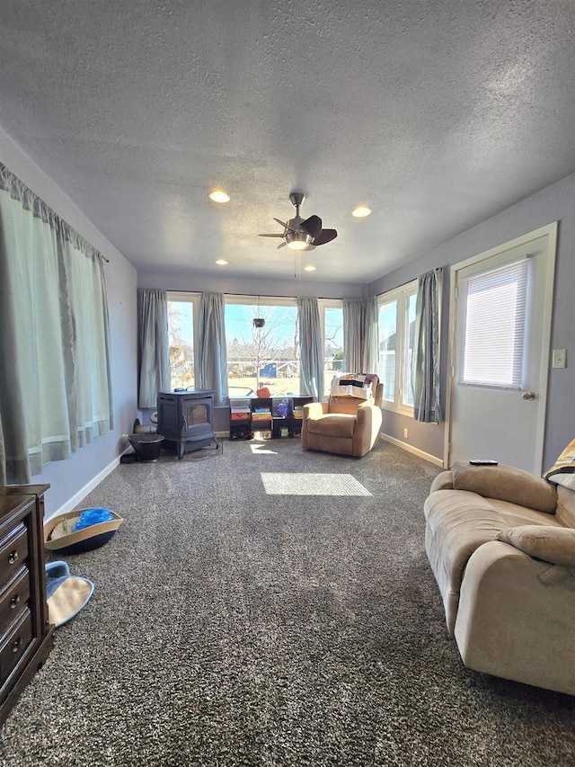 living room featuring a ceiling fan, baseboards, a wood stove, a textured ceiling, and carpet flooring