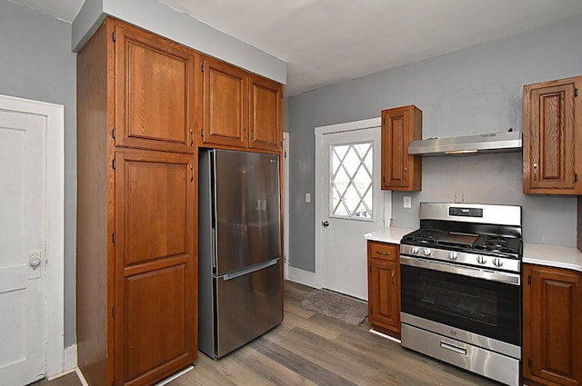 kitchen with under cabinet range hood, light countertops, appliances with stainless steel finishes, wood finished floors, and brown cabinetry