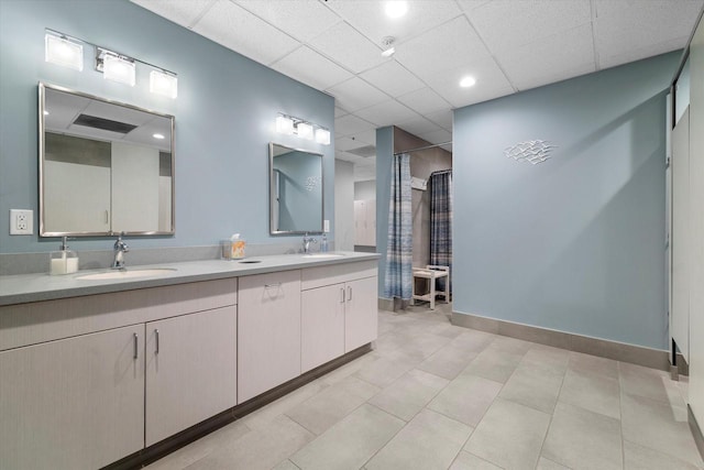 full bath featuring a shower with curtain, baseboards, double vanity, a drop ceiling, and a sink