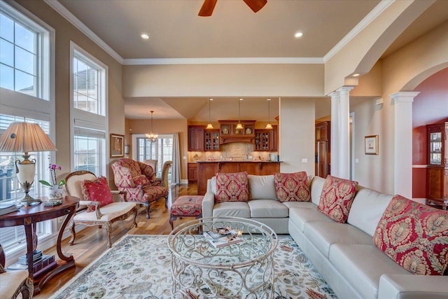 living area featuring light wood-style flooring, a ceiling fan, arched walkways, crown molding, and ornate columns