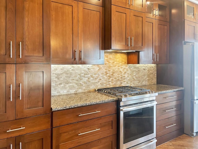 kitchen with tasteful backsplash, glass insert cabinets, light stone countertops, brown cabinetry, and stainless steel appliances