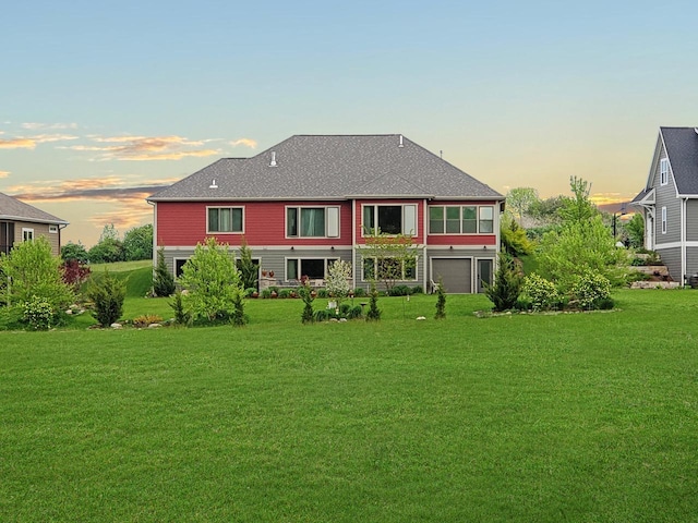 back of property at dusk featuring a lawn, an attached garage, and roof with shingles