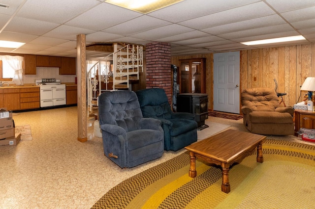 living room featuring a wood stove, a drop ceiling, wooden walls, light floors, and stairs