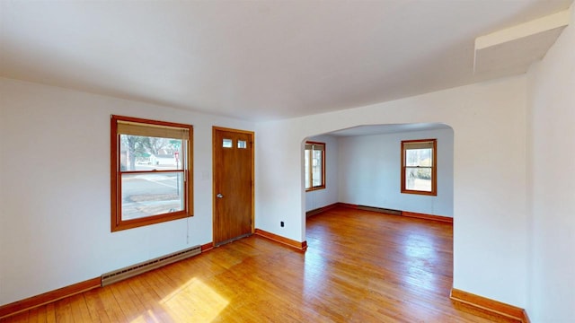 spare room featuring arched walkways, light wood-type flooring, a baseboard heating unit, and a wealth of natural light